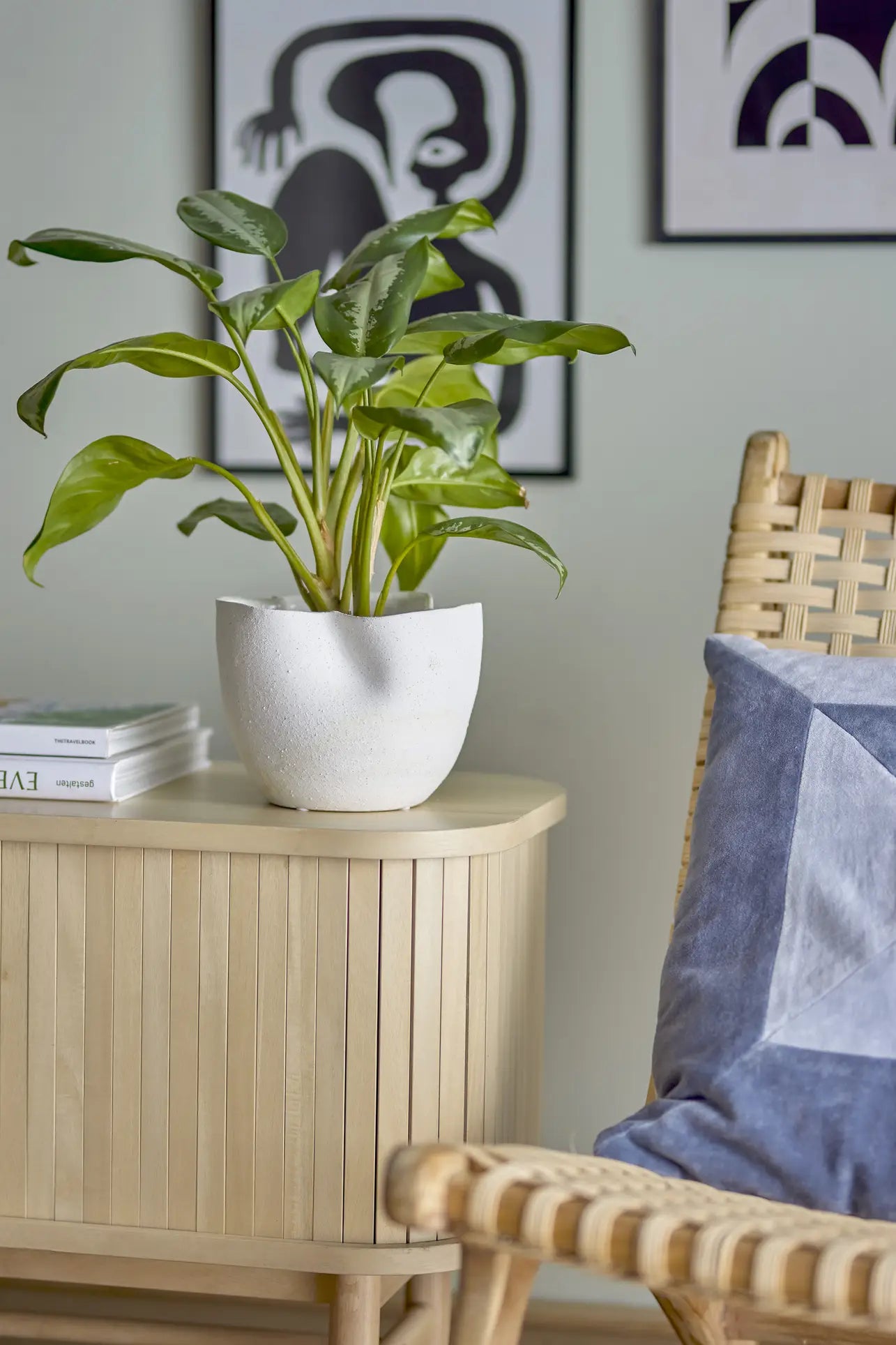 A green plant in the Bloomingville Sanam Flowerpot sits on a light wooden cabinet beside a woven chair with a blue and white pillow, with abstract black-and-white artwork on the wall in the background.