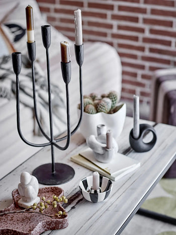 A white coffee table with assorted candles in modern holders, books, decorative objects, and a potted cactus in the Bloomingville Sanam Flowerpot by Bloomingville sits before a striped sofa and red brick wall.