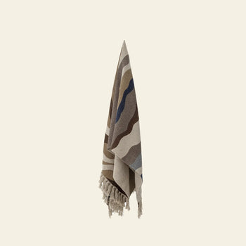 The Bloomingville Stephania Throw by Bloomingville features beige, brown, and blue wavy stripes on recycled cotton, with fringed edges. It hangs vertically against an off-white background.