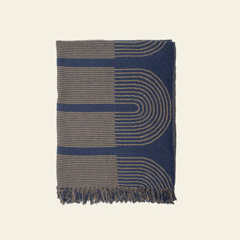 The Bloomingville Tiffanie Throw by Bloomingville is made from soft cotton, featuring a navy blue and tan graphic curved line pattern with fringed edges, shown on a plain light background.