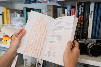 A person holds open Bluetaped Refuse: A Dumpster Diving Craft Handbook by Wong Eng Geng (Bluetaped), standing before a bookshelf filled with books, a camera, and upcycled objects made from furniture waste.