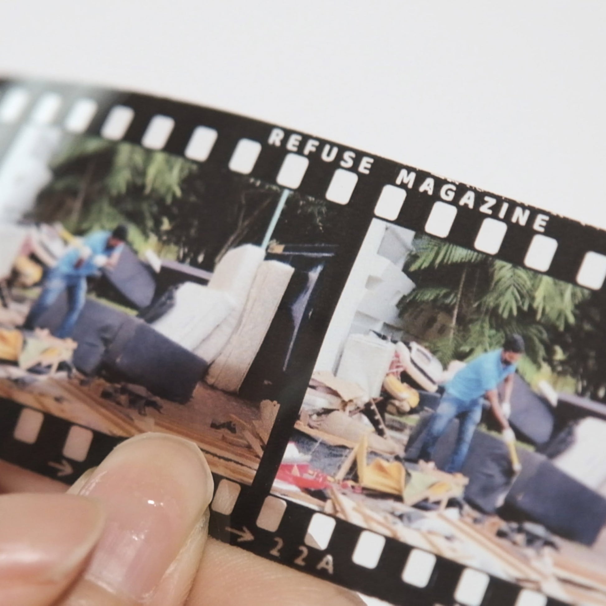 A close-up of a hand holding a film strip labeled Bluetaped Refuse: A Dumpster Diving Craft Handbook by Wong Eng Geng from Bluetaped, featuring two images of someone moving large items among piles of furniture waste outdoors.