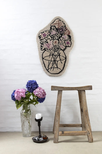 A rustic wooden stool sits by a glass vase of purple and pink hydrangeas; above, the Bongusta Autumn Flowers Rug by Bongusta, crafted from 100% wool, displays a floral vase design against a white brick wall.