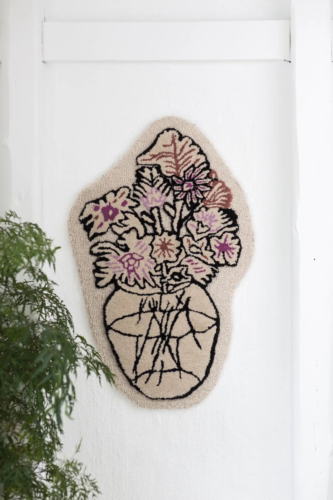 The Bongusta Autumn Flowers Rug by Bongusta is a handcrafted 100% wool wall hanging featuring abstract pink and purple flowers in a vase, outlined in black—perfect on a white wall, especially paired with a leafy green plant.
