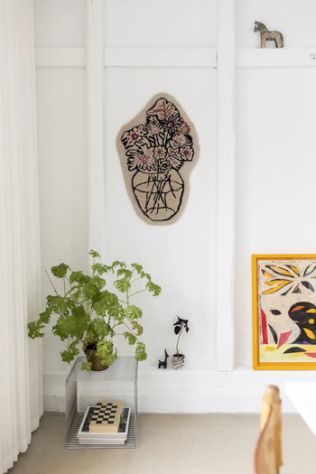 A minimalist room with white walls features the Bongusta Autumn Flowers Rug, a handcrafted 100% wool piece by Bongusta, a floral wall hanging, green potted plant on a clear side table with chessboard, and framed art with yellow accents.
