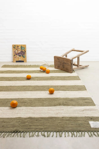 The Bongusta Chindi Rug (140x200) by Bongusta, featuring vibrant stripes and handcrafted in India, lies in a minimalist room with scattered oranges, a tipped-over wooden stool, clear glass, and a small framed picture against a white wall.