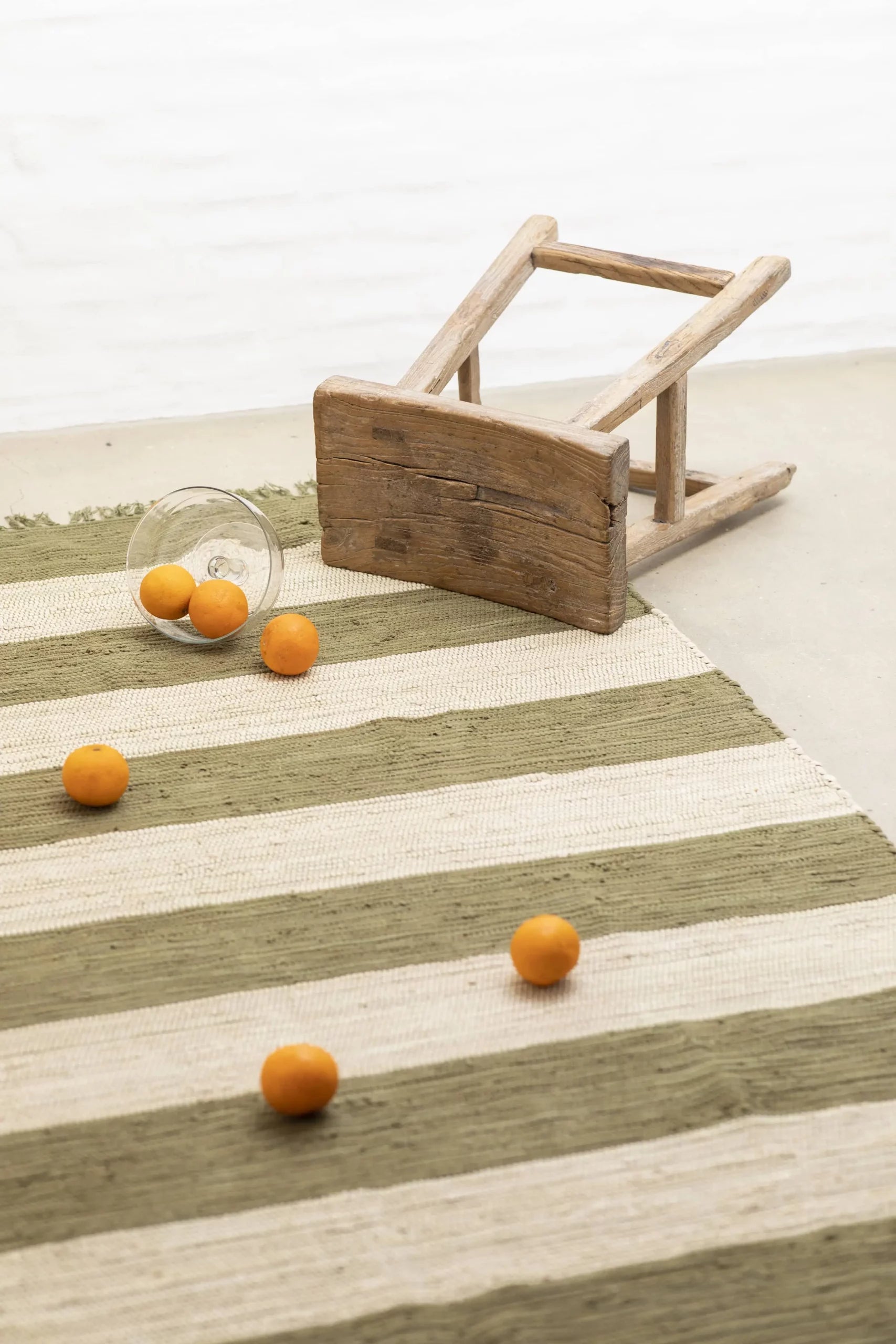 A small wooden stool lies tipped over on the Bongusta Chindi Rug (140x200) by Bongusta, handcrafted in India. Orange fruits and a glass are scattered across the vibrant beige and green rug.