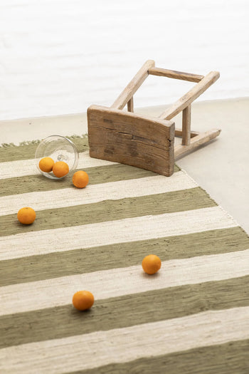 A small wooden stool lies tipped over on the Bongusta Chindi Rug (140x200) by Bongusta, handcrafted in India. Orange fruits and a glass are scattered across the vibrant beige and green rug.