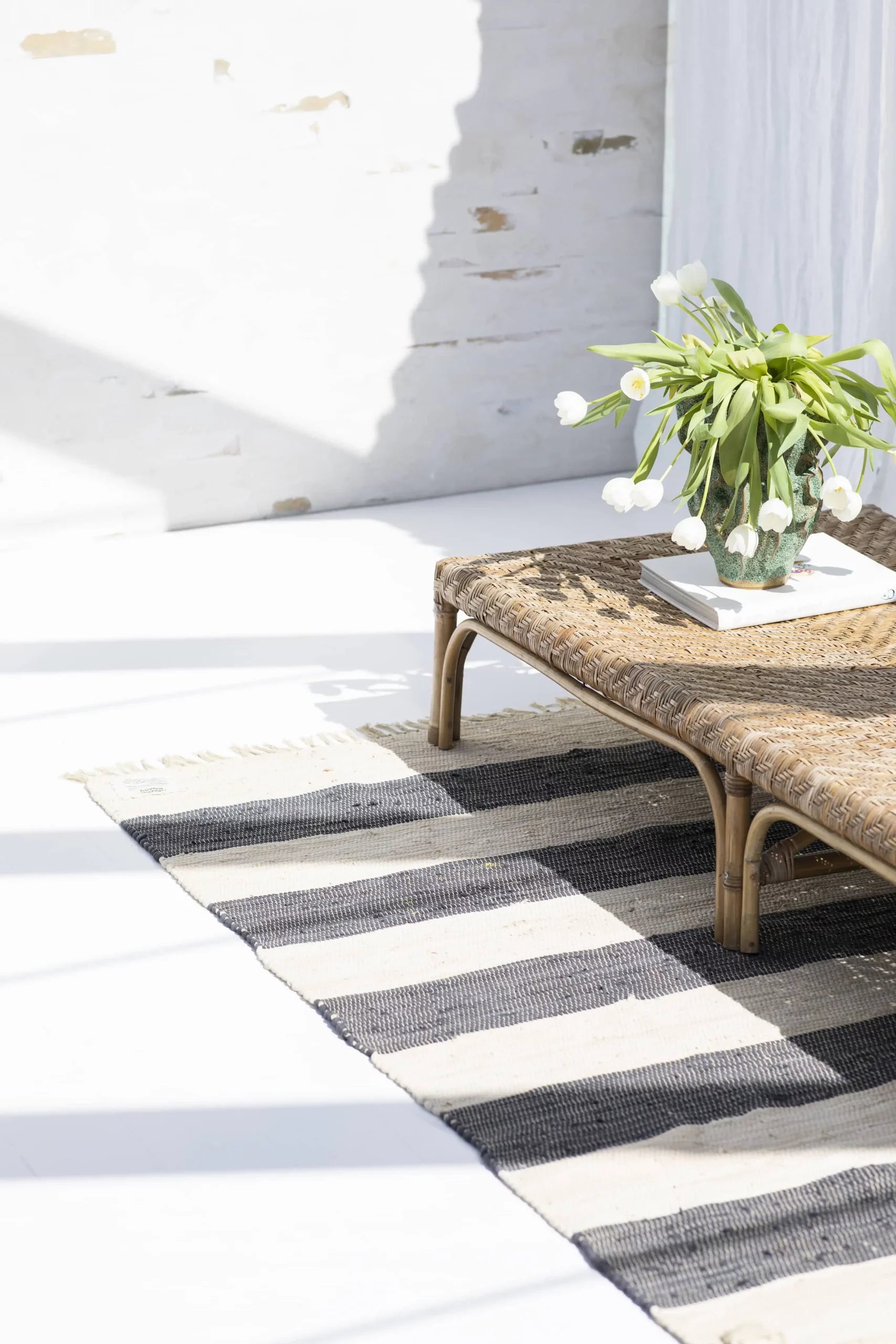 A wicker coffee table with a vase of white tulips and a book sits on the Bongusta Chindi Rug (140x200) by Bongusta, handcrafted in India. Sunlight streams into the bright room, casting windowpane shadows across white walls and floor.