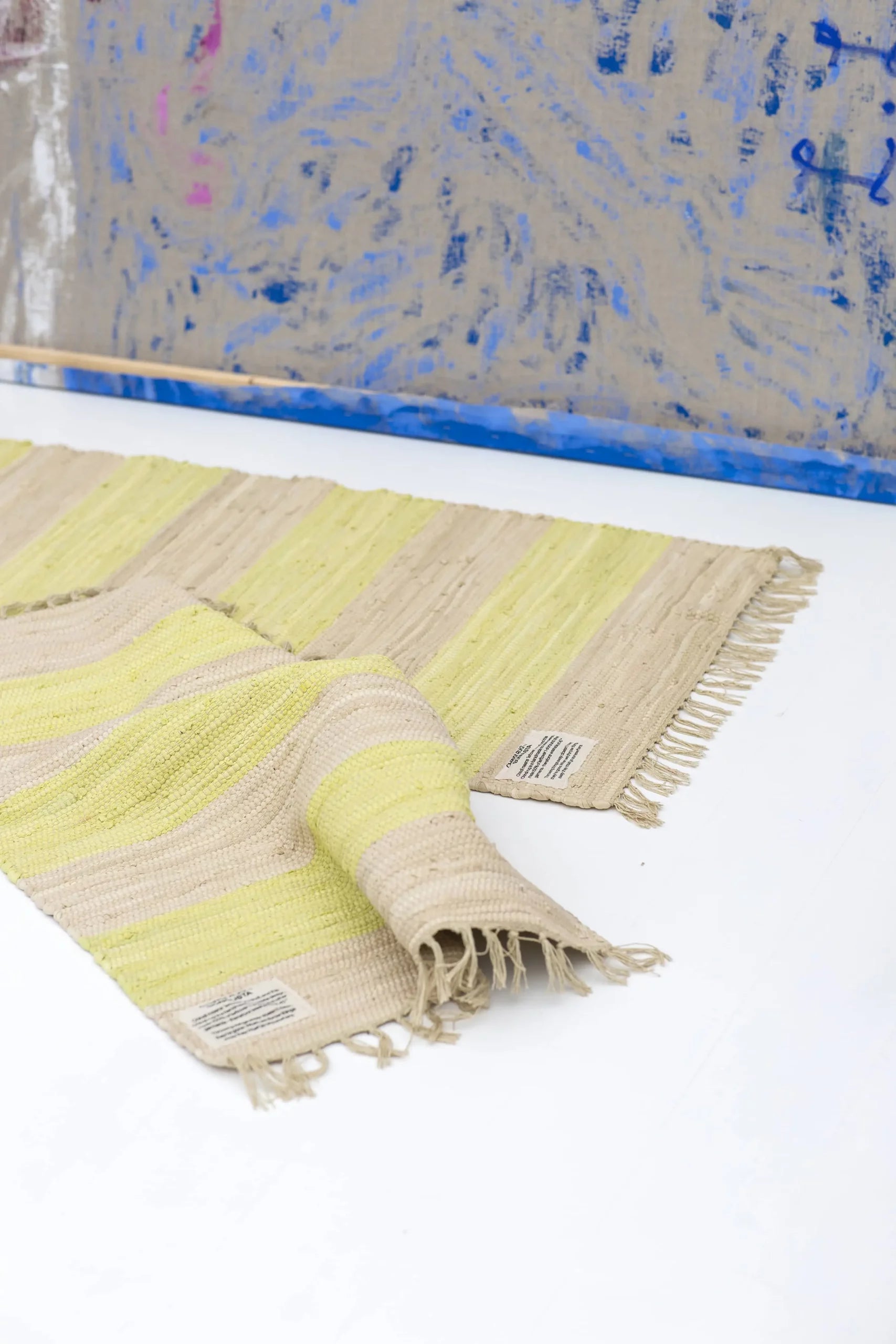 The Bongusta Chindi Rug (45x60) by Bongusta is handcrafted in India from surplus cotton, featuring beige and yellow stripes with fringed edges. It has a sewn label on one corner and is pictured partly folded on a white floor before an abstract wall.