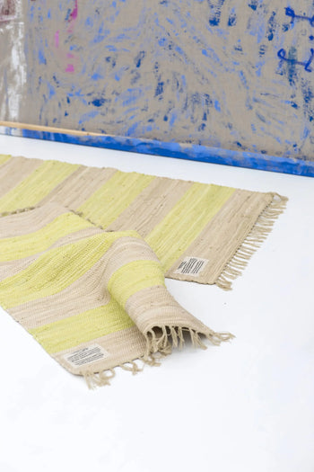 The Bongusta Chindi Rug (45x60) by Bongusta is handcrafted in India from surplus cotton, featuring beige and yellow stripes with fringed edges. It has a sewn label on one corner and is pictured partly folded on a white floor before an abstract wall.