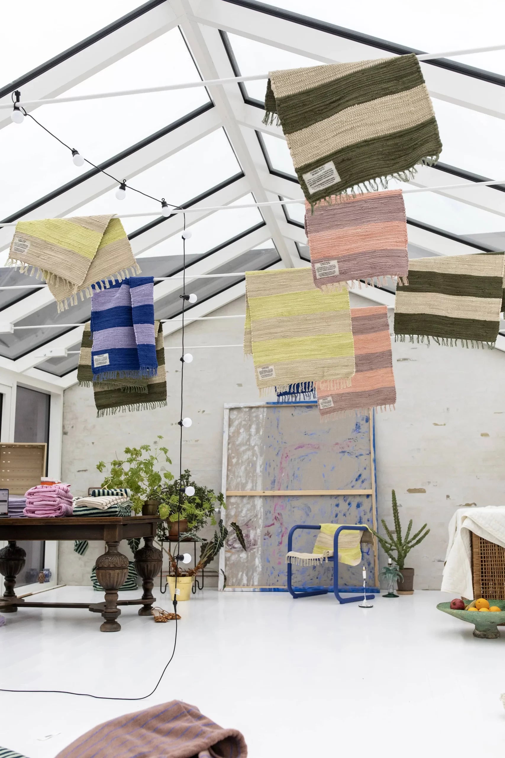 Bongusta Chindi Rugs (45x60), handcrafted in India from surplus cotton by Bongusta, hang from a sunlit glass-roofed room featuring a wooden table, blue chair, lush houseplants, and an abstract painting on a white brick wall.