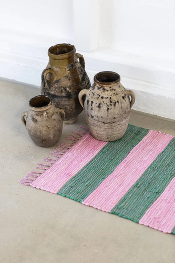 Three rustic earthenware jars with handles sit on a light floor beside the Bongusta Chindi Rug (45x60) by Bongusta, handcrafted in India from surplus cotton with pink and green stripes and fringe. The backdrop is a white wall.