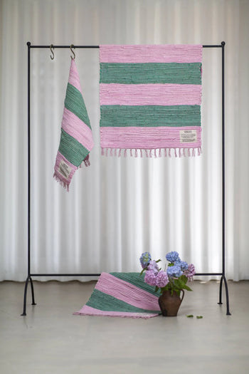 A black metal rack displays two Bongusta Chindi Rugs (45x60) by Bongusta, handcrafted in India from surplus cotton with pink and green stripes; a matching rug is beside a brown vase of hydrangeas. White curtains hang behind.