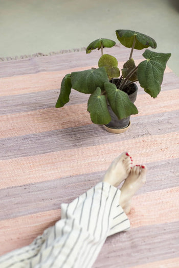 A potted plant with large green leaves sits on the Bongusta Chindi Rug (45x60), handcrafted in India from surplus cotton fabric. At the bottom, a person with painted toenails and striped pants rests their feet near the plant.