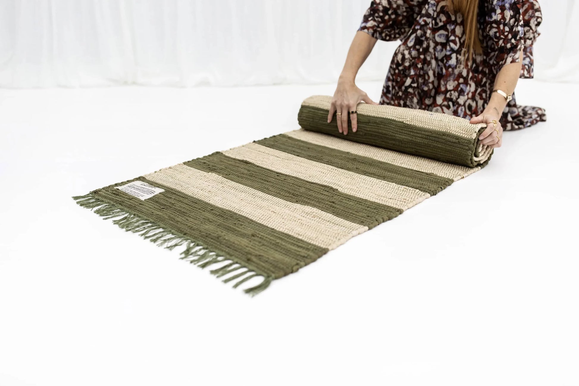 A person in a patterned dress rolls out the Bongusta Chindi Rug (60x120) by Bongusta, featuring green and beige stripes, fringed edges, and handcrafted in India from surplus cotton fabric, on a white floor.