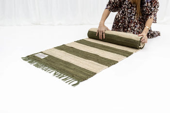 A person in a patterned dress rolls out the Bongusta Chindi Rug (60x120) by Bongusta, featuring green and beige stripes, fringed edges, and handcrafted in India from surplus cotton fabric, on a white floor.