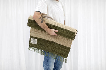 A person in a white shirt and blue jeans holds the Bongusta Chindi Rug (60x120) by Bongusta—handcrafted in India from surplus cotton, two-tone green and beige with tassels and label—standing before a white curtain background.