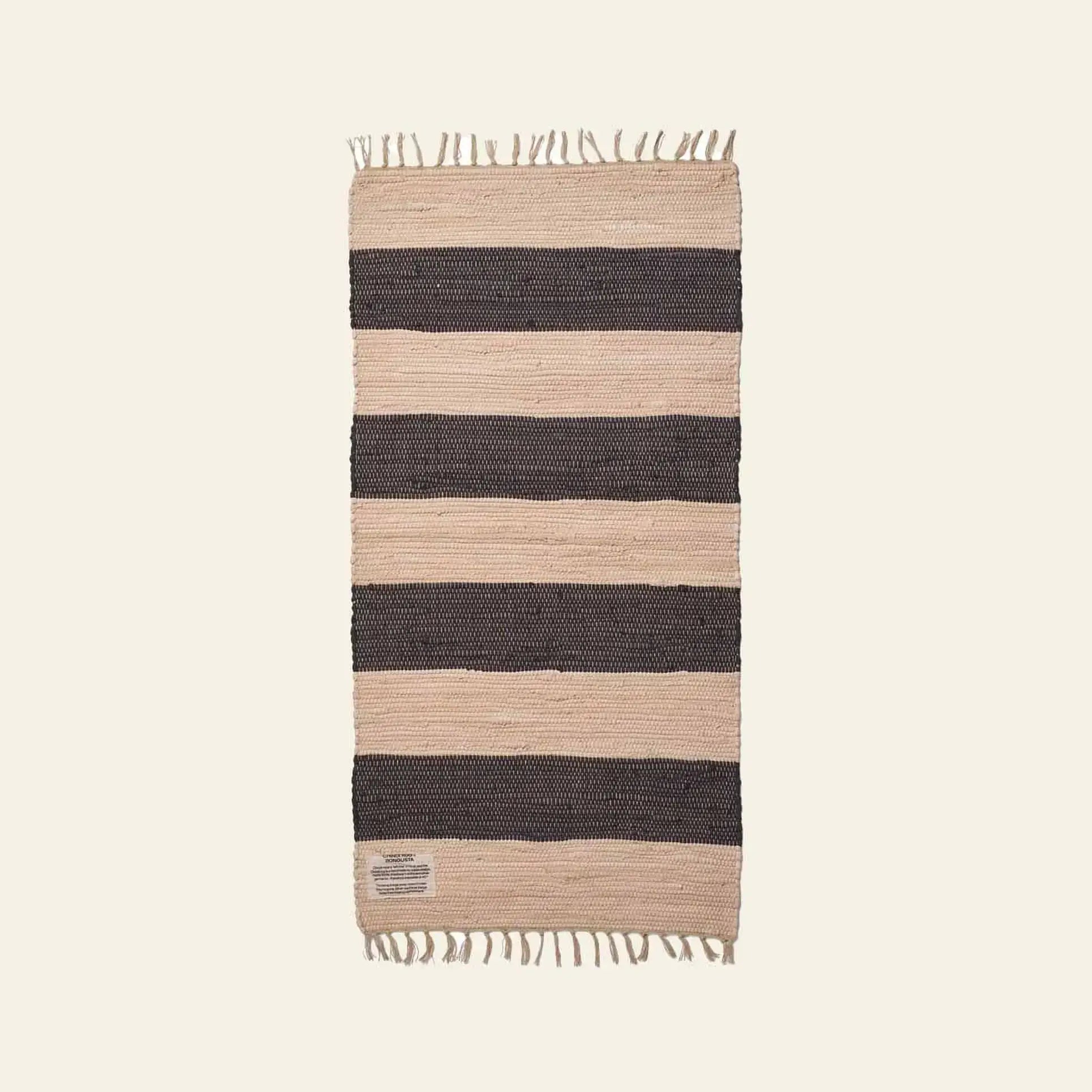The Bongusta Chindi Rug (60x120) by Bongusta is a rectangular rug handcrafted in India from surplus cotton, with wide black and beige stripes and short fringe on the ends, shown against a plain light background.