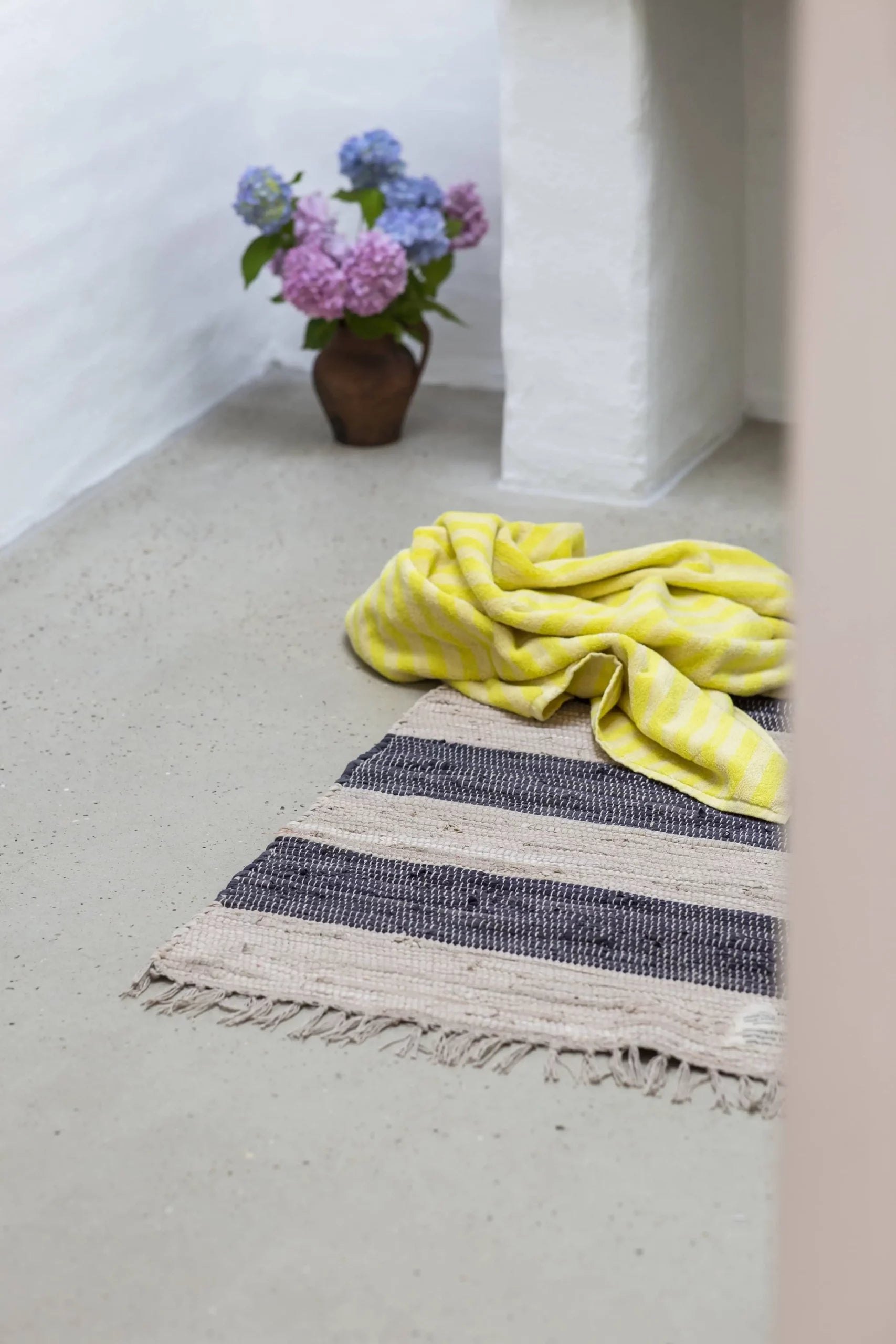 A yellow striped surplus cotton fabric is casually draped on a light gray floor next to a Bongusta Chindi Rug (60x120) by Bongusta. In the background, a brown vase holds pink and purple flowers against a white wall.