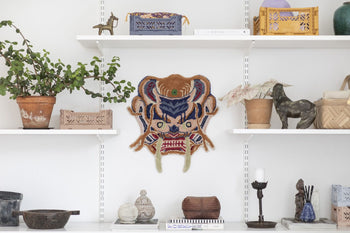 White shelves adorned with potted plants, woven baskets, ceramics, books, and candle holders frame the Bongusta Dragon Face Rug by Bongusta on the central wall, displayed beside a vibrant embroidered mask.