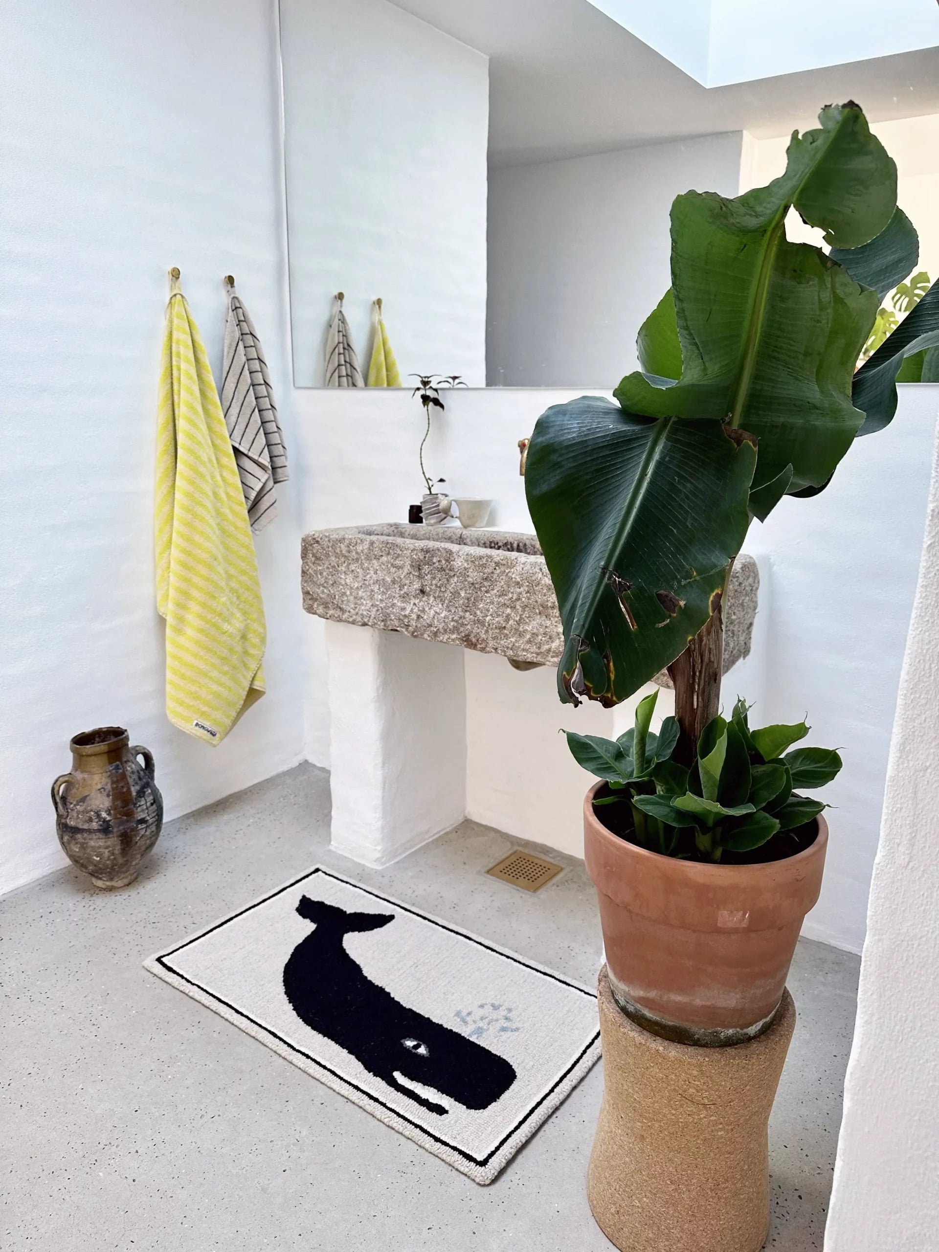 A bright, minimalist bathroom with white walls, stone sink, potted plant, yellow and striped towels, decorative jug, and the Bongusta Frame Rug—a playful alternative to wall decor featuring a whale print—on the floor.