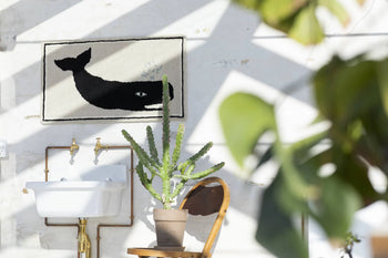 Sunlight streams into a bright room featuring a Bongusta Frame Rug by Bongusta, a white sink, wooden chair with potted cactus, and unique wall decor showing a black whale. Blurred green leaves appear in the foreground.