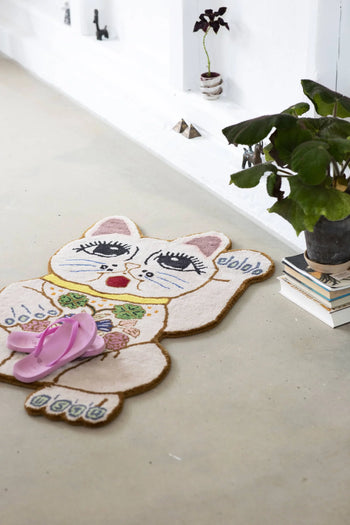 The Bongusta Lucky Rug, hand-tufted from wool, features floral accents on its cat-shaped design. Pink flip-flops rest on it while a potted plant atop stacked books sits nearby against a white wall.