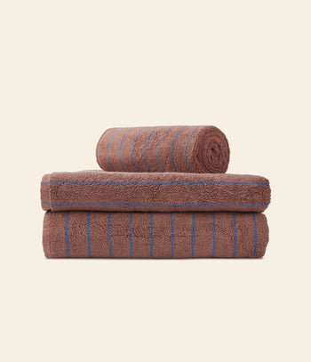 Three brown Bongusta Naram Bath Towels with thin blue stripes are stacked neatly against a light background. Made from Oeko-Tex combed cotton, one luxury towel is rolled on top of two folded towels by Bongusta.