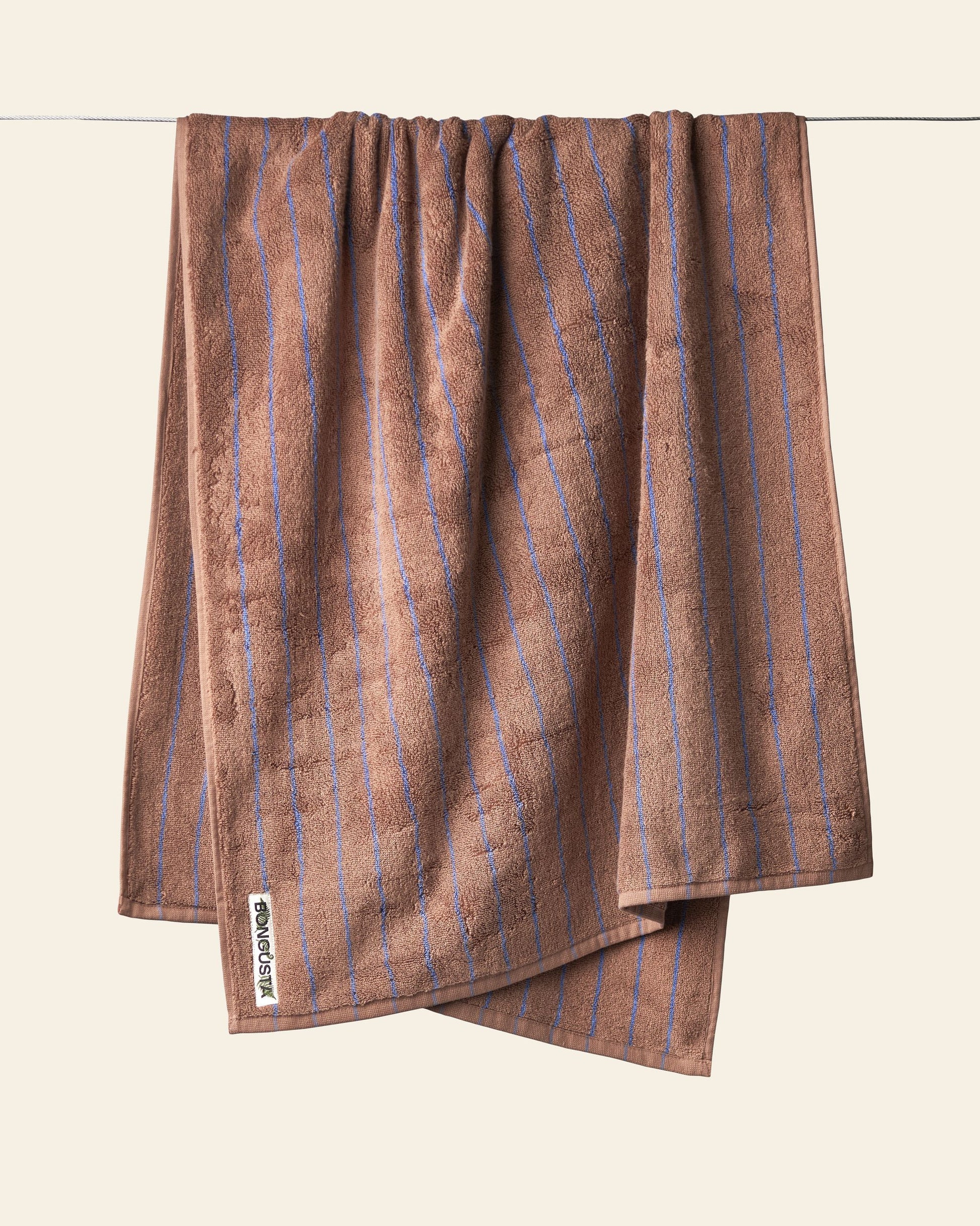 A Bongusta Naram Bath Towel with thin blue vertical stripes hangs on a clothesline against a beige background. Crafted from Oeko-Tex combed cotton, its Bongusta luxury tag is visible, promising style and comfort.