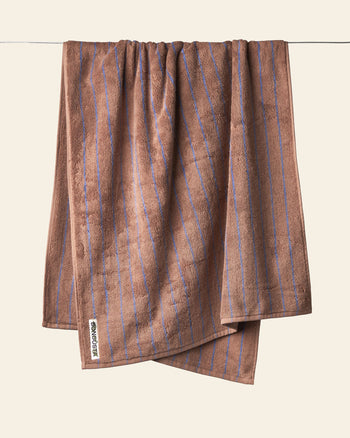 A Bongusta Naram Bath Towel with thin blue vertical stripes hangs on a clothesline against a beige background. Crafted from Oeko-Tex combed cotton, its Bongusta luxury tag is visible, promising style and comfort.