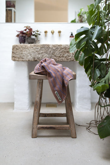 A rustic wooden stool with a folded Bongusta Naram Bath Towel, made from Oeko-Tex combed cotton by Bongusta, sits before a stone sink in a bright bathroom filled with green potted plants.
