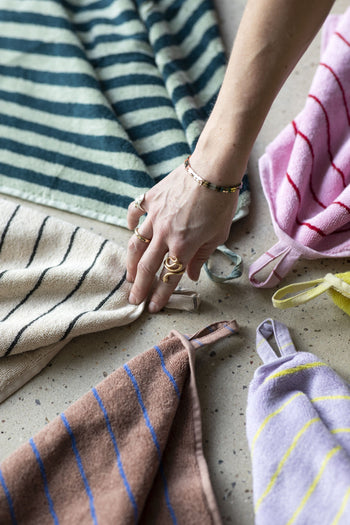 A hand with rings and a bracelet touches a beige Bongusta Naram Bath Towel with black stripes, displayed among other colorful striped Bongusta towels on a speckled surface, showcasing luxury bath towel elegance.