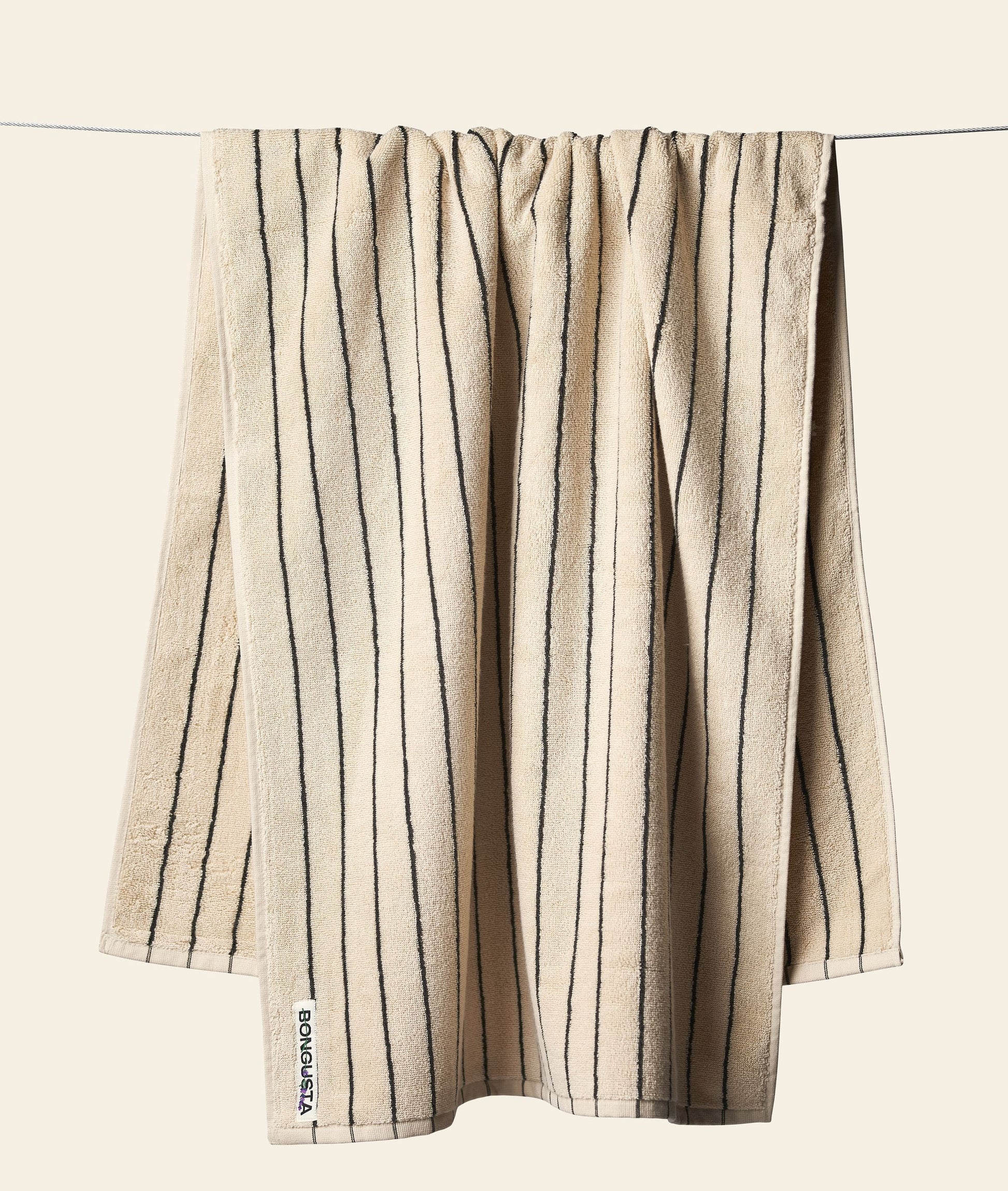 A beige Bongusta Naram Bath Towel made from Oeko-Tex combed cotton with vertical dark stripes hangs on a clothesline against a plain light background. A small white tag with black text is on the bottom left corner. Brand: Bongusta.
