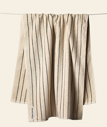 A beige Bongusta Naram Bath Towel made from Oeko-Tex combed cotton with vertical dark stripes hangs on a clothesline against a plain light background. A small white tag with black text is on the bottom left corner. Brand: Bongusta.