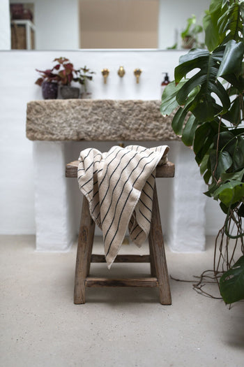 A Bongusta Naram Bath Towel is draped over a rustic wooden stool in front of a stone sink, surrounded by green plants in a minimalist bathroom.