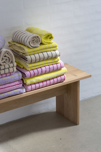 Stacks of Bongusta Naram Bath Towels in yellow, beige, pink, and purple—crafted from Oeko-Tex combed cotton—are neatly arranged on a wooden bench against a white brick wall for an elegant bath upgrade.