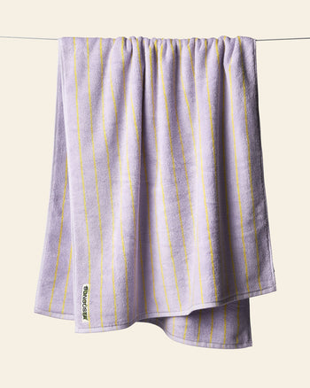 A Bongusta Naram Bath Towel in lavender with thin yellow stripes hangs on a clothesline against a light beige background. Made from Oeko-Tex combed cotton, this luxury towel by Bongusta displays a small white tag in one corner.