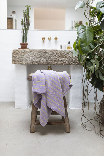 A rustic bathroom features a Bongusta Naram Bath Towel by Bongusta in purple with yellow stripes, draped on a wooden stool beside a stone sink, brass faucets, and potted plants for a minimalist, refined natural decor.