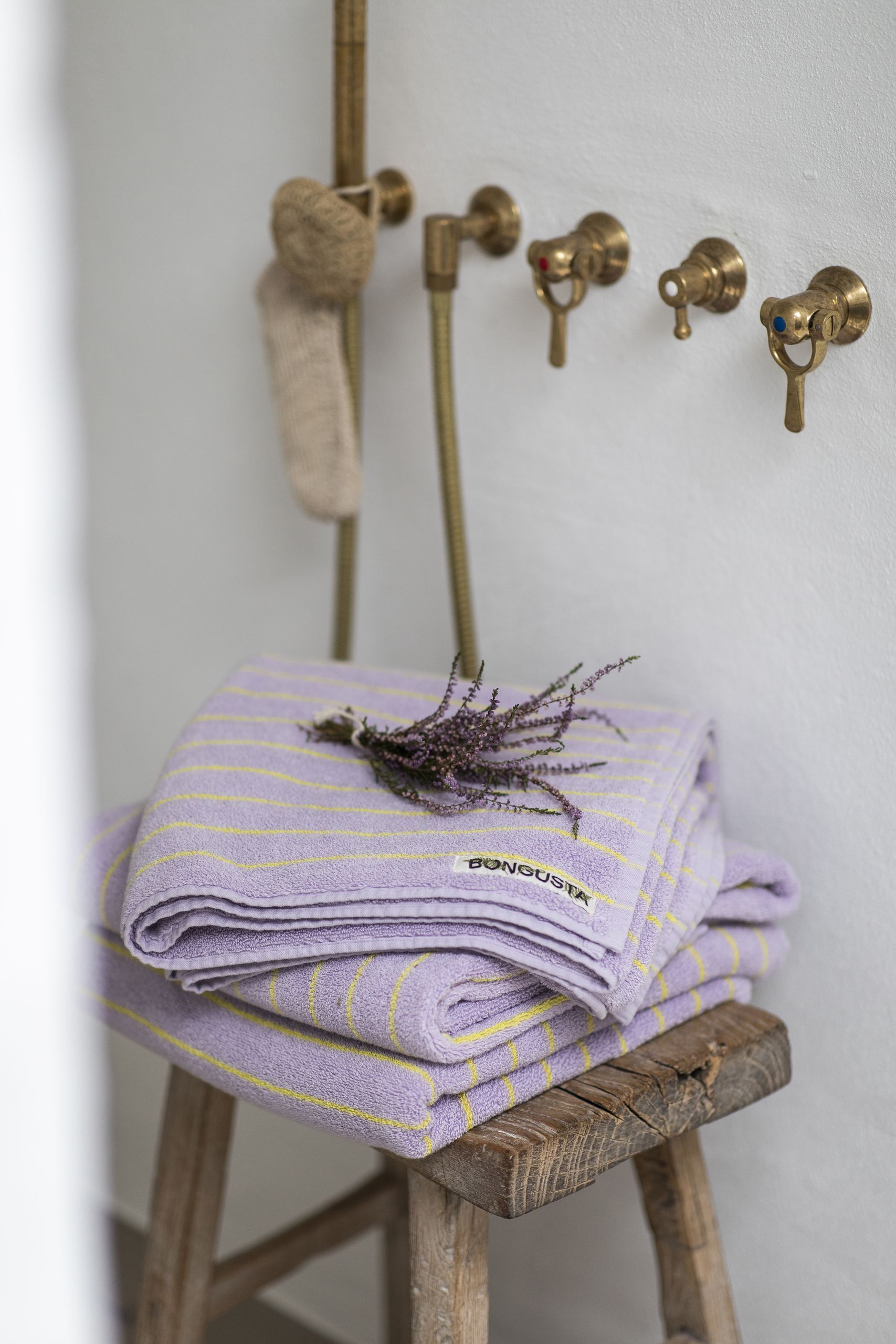 Two folded Bongusta Naram Bath Towels in lavender stripes rest on a wooden stool topped with lavender. Crafted from Oeko-Tex combed cotton, these luxury Bongusta towels sit near brass fixtures and a scrubber by a white wall.