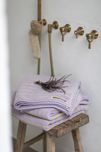 Two folded Bongusta Naram Bath Towels in lavender stripes rest on a wooden stool topped with lavender. Crafted from Oeko-Tex combed cotton, these luxury Bongusta towels sit near brass fixtures and a scrubber by a white wall.