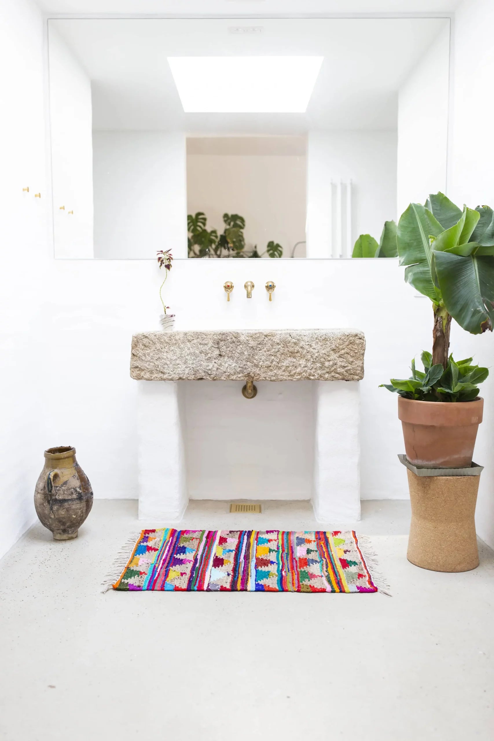 A minimalist bathroom features a stone sink, gold faucet, large mirror, potted plant on a stand, rustic vase, and the Bongusta Pyramid Chindi Rug (60x90) by Bongusta—its recycled cotton brightens the white floor.