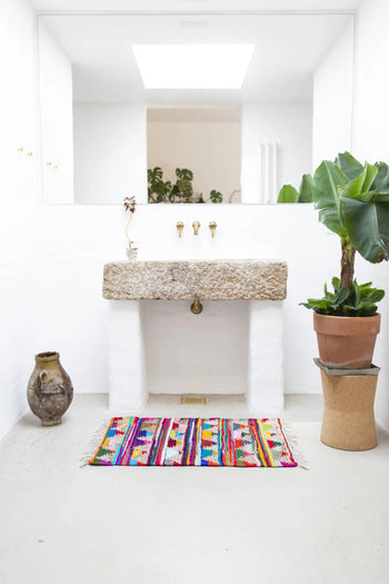 A minimalist bathroom features a stone sink, gold faucet, large mirror, potted plant on a stand, rustic vase, and the Bongusta Pyramid Chindi Rug (60x90) by Bongusta—its recycled cotton brightens the white floor.
