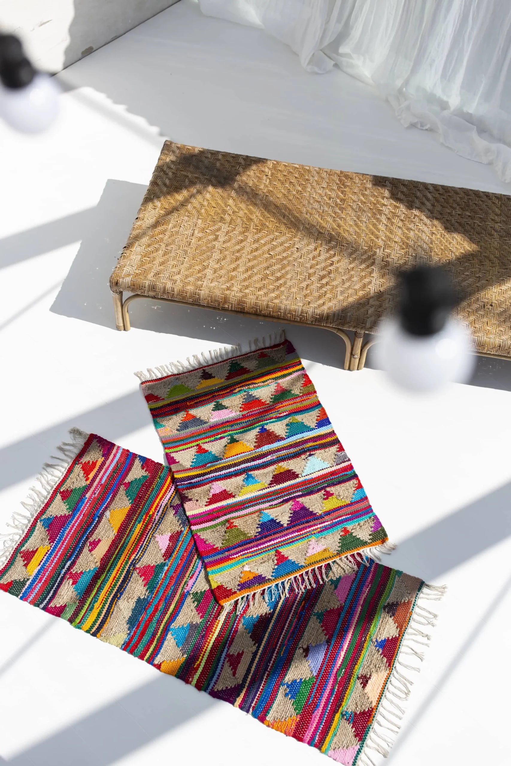 A woven rattan bench and two Bongusta Pyramid Chindi Rugs (60x90) with geometric patterns rest on a white floor, as shadows and light bulbs create striking patterns and cotton fabric and jute yarn add textured vibrancy.