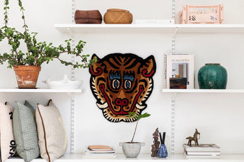 White shelves showcase decor like a potted plant, baskets, books, pillows, and sculptures. Centered between them is the Bongusta Tiger Face Rug by Bongusta—a vibrant handwoven piece that adds striking color as a wall hanging.