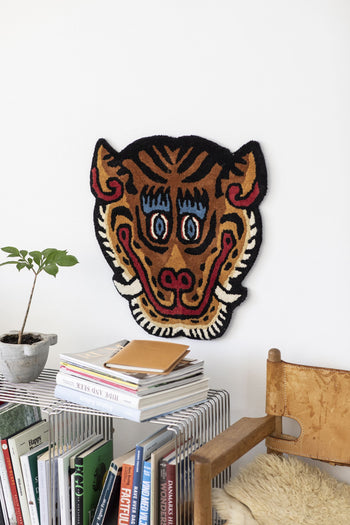 The Bongusta Tiger Face Rug by Bongusta, a vibrant textured wall hanging shaped like a stylized tiger face with bold blue eyes and pointed ears, is shown above a small table with books, a potted plant, and a wooden chair nearby.