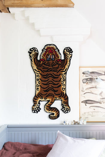 A Bongusta Tiger Rug, Baby by Bongusta is mounted on a white wall under a staircase next to a framed animal illustration, creating a jungle vibe. Below, part of a gray bed with a red blanket can be seen.