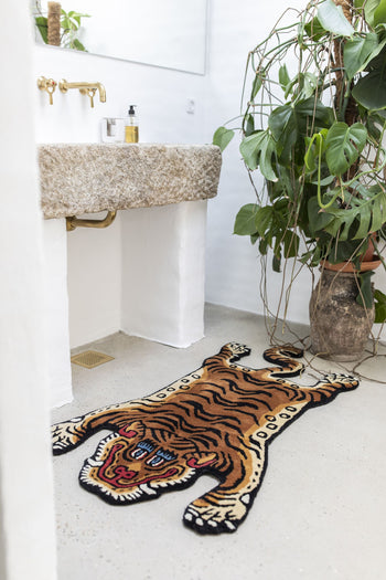 A bathroom with a stone sink and gold faucet features a potted plant and the Bongusta Tiger Rug, Small by Bongusta, showcasing its playful tiger design on the light-colored floor.