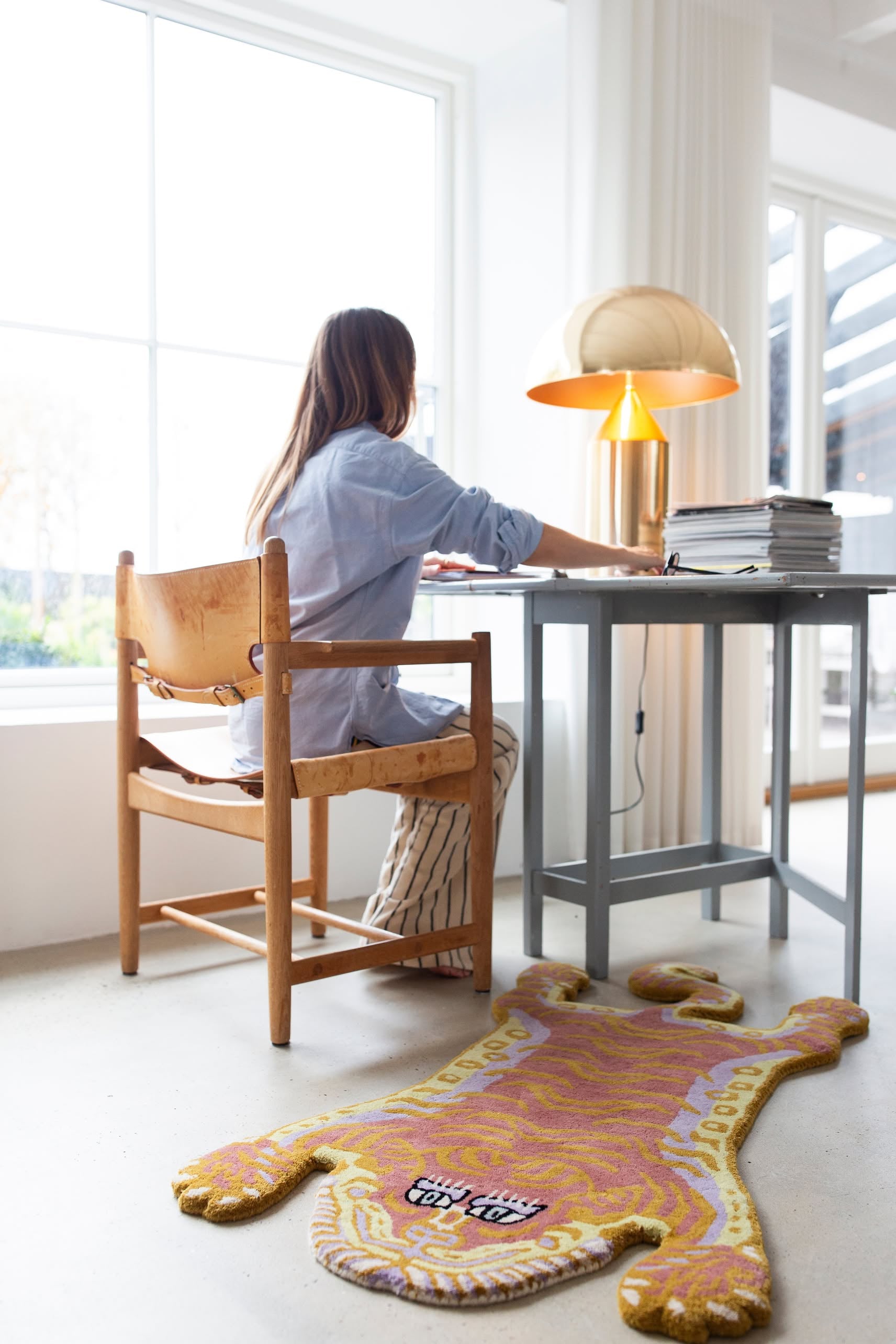 A woman works at a desk in a bright, windowed room. Nearby, stacks of papers, a gold lamp, and the Bongusta Tigress Rug, Small—by Bongusta with its playful tiger motif—add character to the space.