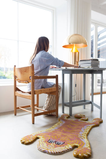 A woman works at a desk in a bright, windowed room. Nearby, stacks of papers, a gold lamp, and the Bongusta Tigress Rug, Small—by Bongusta with its playful tiger motif—add character to the space.